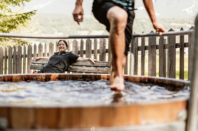 A woman is sitting on a bench in front of a wooden barrel in which a person is walking through water with his feet.