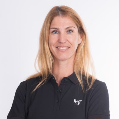 Frau mit langen blonden Haaren in einem dunkelblauen Poloshirt mit dem Logo "IMST". Sie lächelt und hat die Arme verschränkt. Heller Hintergrund.