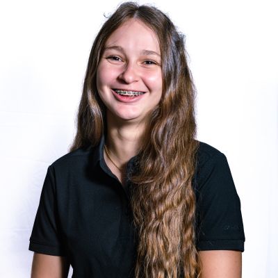 Eine junge Frau mit langen, lockigen Haaren lächelt freundlich in die Kamera. Sie trägt ein schwarzes Poloshirt und hat einen entspannten Gesichtsausdruck. Der Hintergrund ist hell.