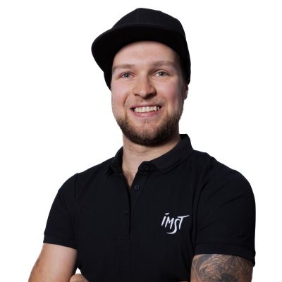 Ein junger Mann steht lächelnd mit verschränkten Armen vor einem weißen Hintergrund. Er trägt ein schwarzes Poloshirt mit einem aufgedruckten Logo.