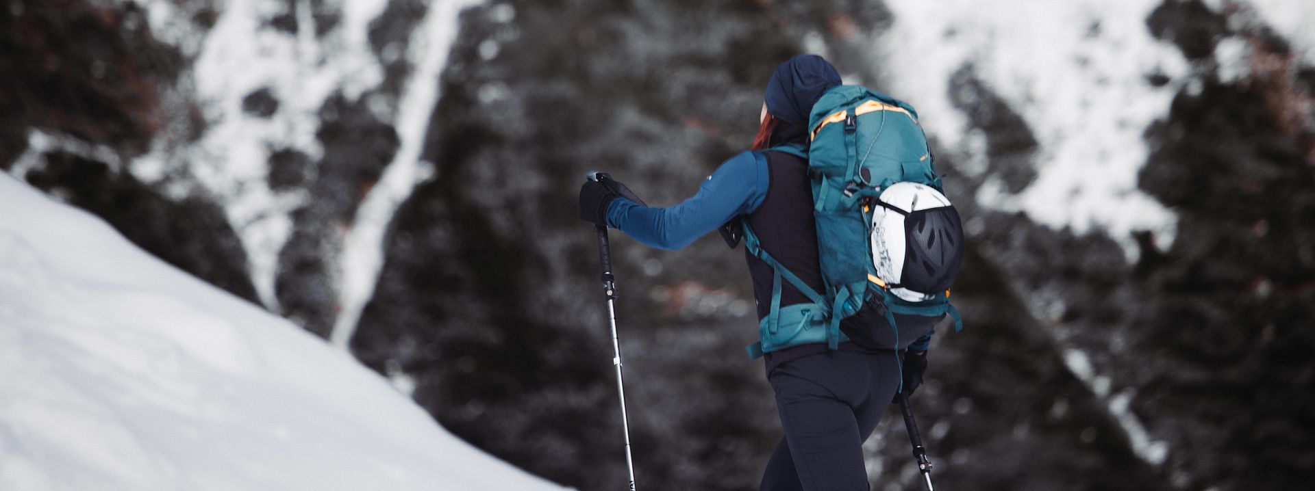 Eine Person mit Rucksack erklimmt einen verschneiten Hang in den Bergen. Der Hintergrund zeigt schneebedeckte Felsen und eine beeindruckende, winterliche Landschaft.