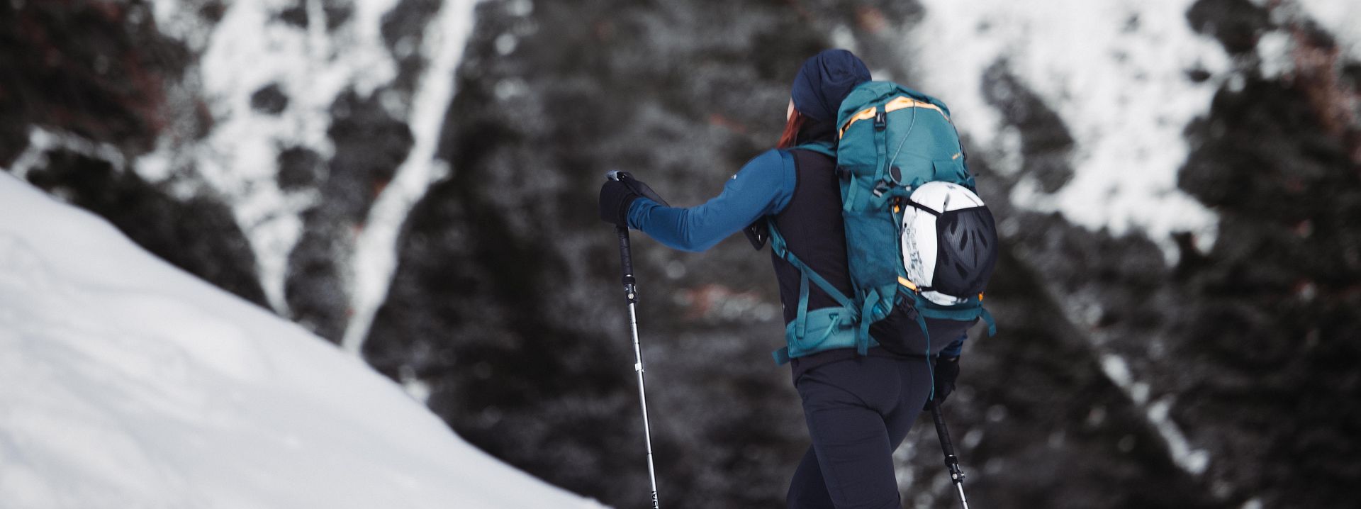 Eine Person mit Rucksack erklimmt einen verschneiten Hang in den Bergen. Der Hintergrund zeigt schneebedeckte Felsen und eine beeindruckende, winterliche Landschaft.