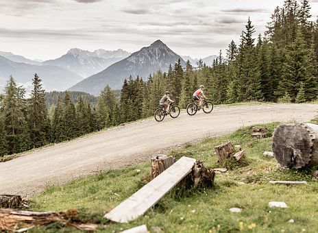 Es sind zwei Personen zu sehen, die mit dem Rad einen Weg in den Bergen entlang fahen.