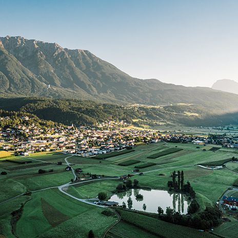 Ein malerisches Dorf liegt in einem grünen Tal unter hohen Bergen. Die Morgensonne beleuchtet sanft das Tal, während Schatten die Bergseite bedecken.
