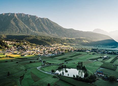 Ein malerisches Dorf liegt in einem grünen Tal unter hohen Bergen. Die Morgensonne beleuchtet sanft das Tal, während Schatten die Bergseite bedecken.