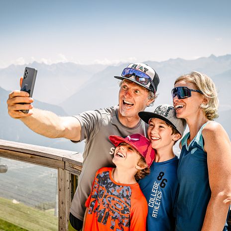 Eine Familie macht ein Selfie an einem Aussichtspunkt vor einer Bergkulisse.