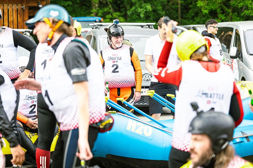 Eine Gruppe von Menschen in Schutzkleidung und Helmen bereitet sich auf ein Rafting-Abenteuer vor. Im Hintergrund sind Boote und Fahrzeuge zu sehen.