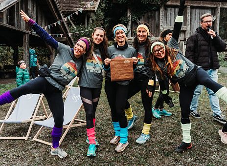 Fünf Frauen in sportlicher Kleidung und bunten, knielangen Socken posieren fröhlich im Freien. Sie halten ein Schild in der Mitte, umgeben von Holzgebäuden und Feierlichkeiten.