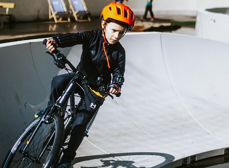 Ein Junge fährt mit einem Fahrrad in einem Pumptrack. Er trägt einen orangefarbenen Helm und sportliche Kleidung. Im Hintergrund sind andere Kinder und Sitzgelegenheiten zu sehen.