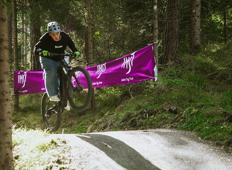 Ein Radfahrer springt mit seinem Mountainbike über einen kleinen Hügel in einem Wald. Im Hintergrund sieht man ein großes, lila Banner, das sich zwischen den Bäumen abhebt.