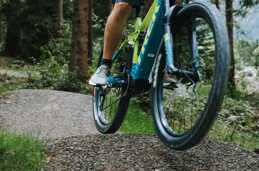 Ein Radfahrer auf einem Mountainbike fährt durch einen Wald. Die Beine des Fahrers sind sichtbar, während er sich auf das Terrain konzentriert. Um ihn herum sind grüne Bäume.