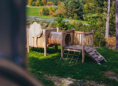 Ein Spielplatz im Freien mit einem Holzspielgerät. Es hat ein Tunnel, eine Kletterwand und eine Rutschbahn. Umgeben von grünen Wiesen und Bäumen im Hintergrund.