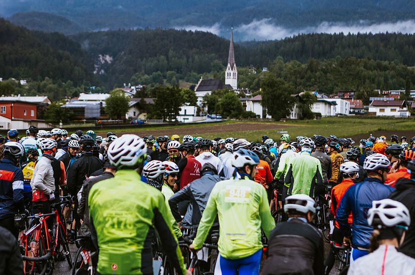 Eine große Gruppe von Radfahrern mit verschiedenen Trikots und Helmen versammelt sich in einer offenen Landschaft. Im Hintergrund ist ein Dorf mit einer Kirche und bewaldeten Hügeln zu sehen.
