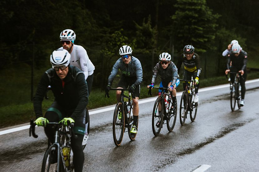 Eine Gruppe von sechs Radfahrern fährt auf einer regnerischen Straße. Alle tragen Helme und sportliche Kleidung. Die Umgebung ist grün, mit Bäumen im Hintergrund.
