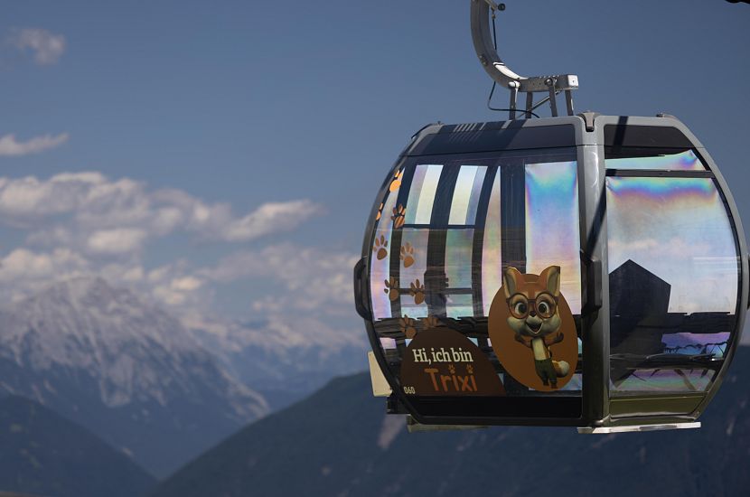 Eine farbenfrohe Gondel schwebt über eine majestätische Berglandschaft. Im Vordergrund sieht man einen niedlichen, cartoonhaften Tier-Charakter auf der Gondel, der lächelt.