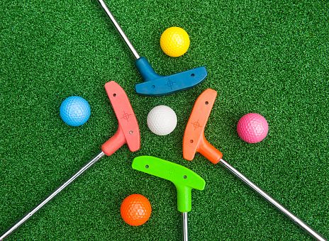 Auf einem grünen, gut gepflegten Golfplatz liegen verschiedene buntfarbige Golfbälle und Putter in Form einer Blume angeordnet. Die Szene strahlt Spaß und Spiel aus.