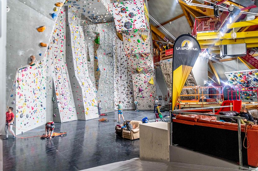 Eine moderne Kletterhalle mit hohen, bunten Kletterwänden. Mehrere Personen klettern oder bereiten sich vor. Eine Flagge mit Logo ist sichtbar. Helle Beleuchtung.