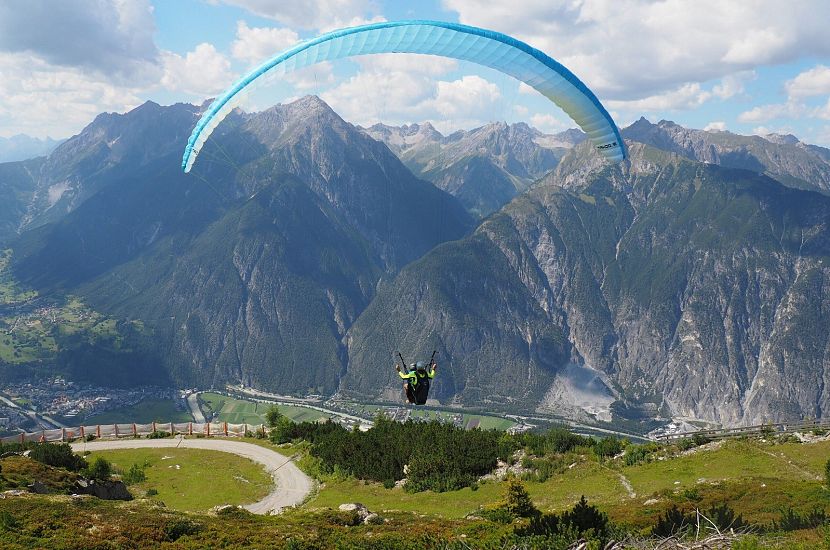 Ein Gleitschirmflieger schwebt hoch über einer malerischen Berglandschaft mit grünen Wiesen und felsigen Gipfeln unter einem strahlend blauen Himmel.
