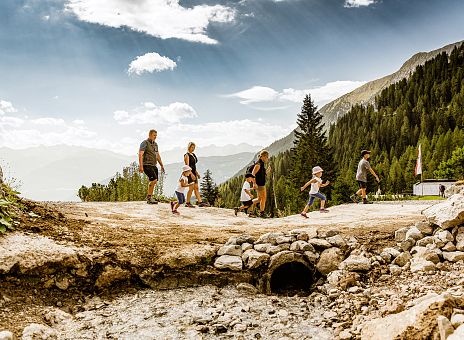 Eine Gruppe von Menschen wandert auf einem Bergpfad in einer malerischen Alpenlandschaft. Der Himmel ist klar, und im Hintergrund sind Tannen und Berggipfel zu sehen.